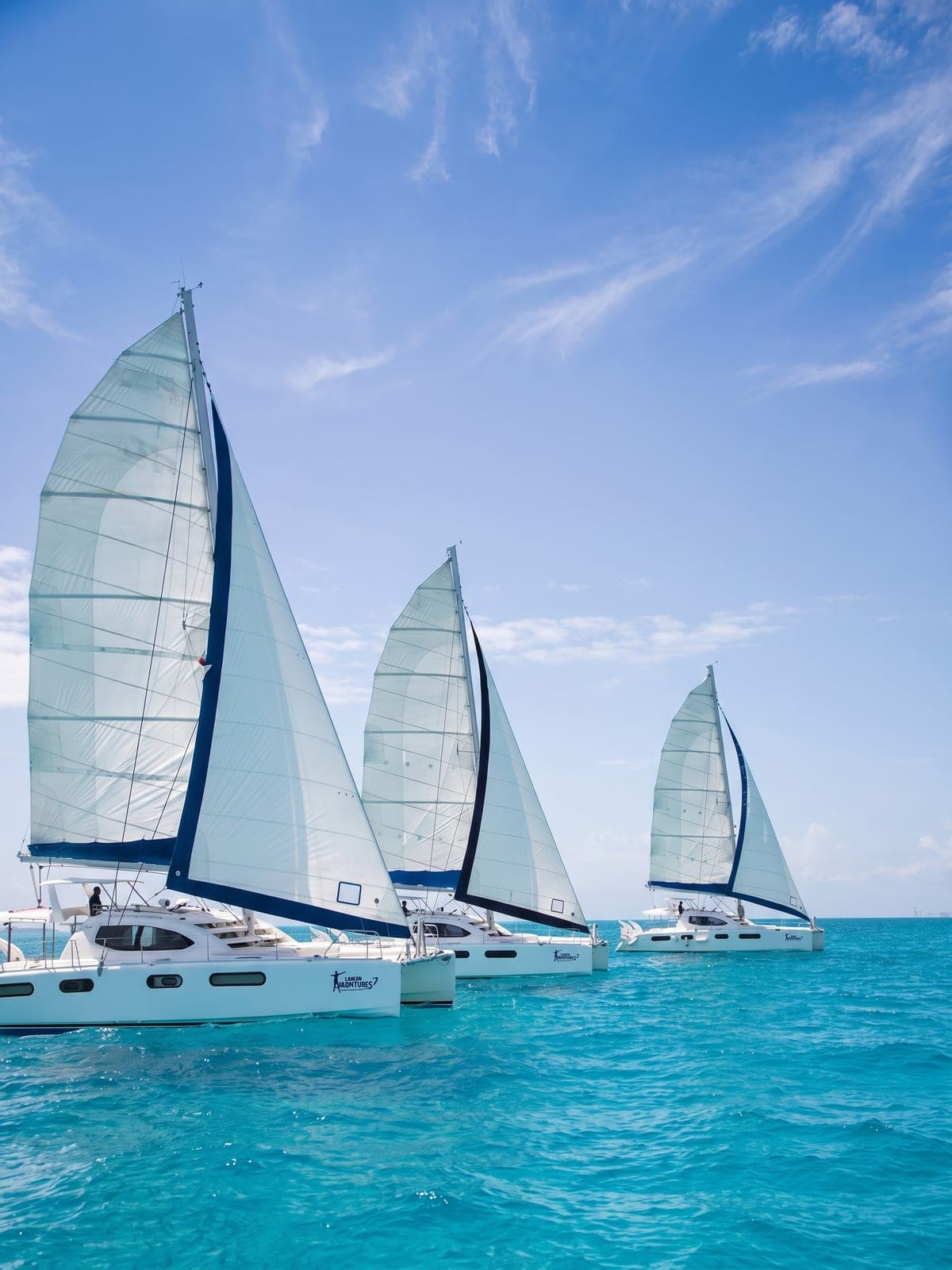 Several sailing catamarans cruising together over turquoise water toward Isla Mujeres.
