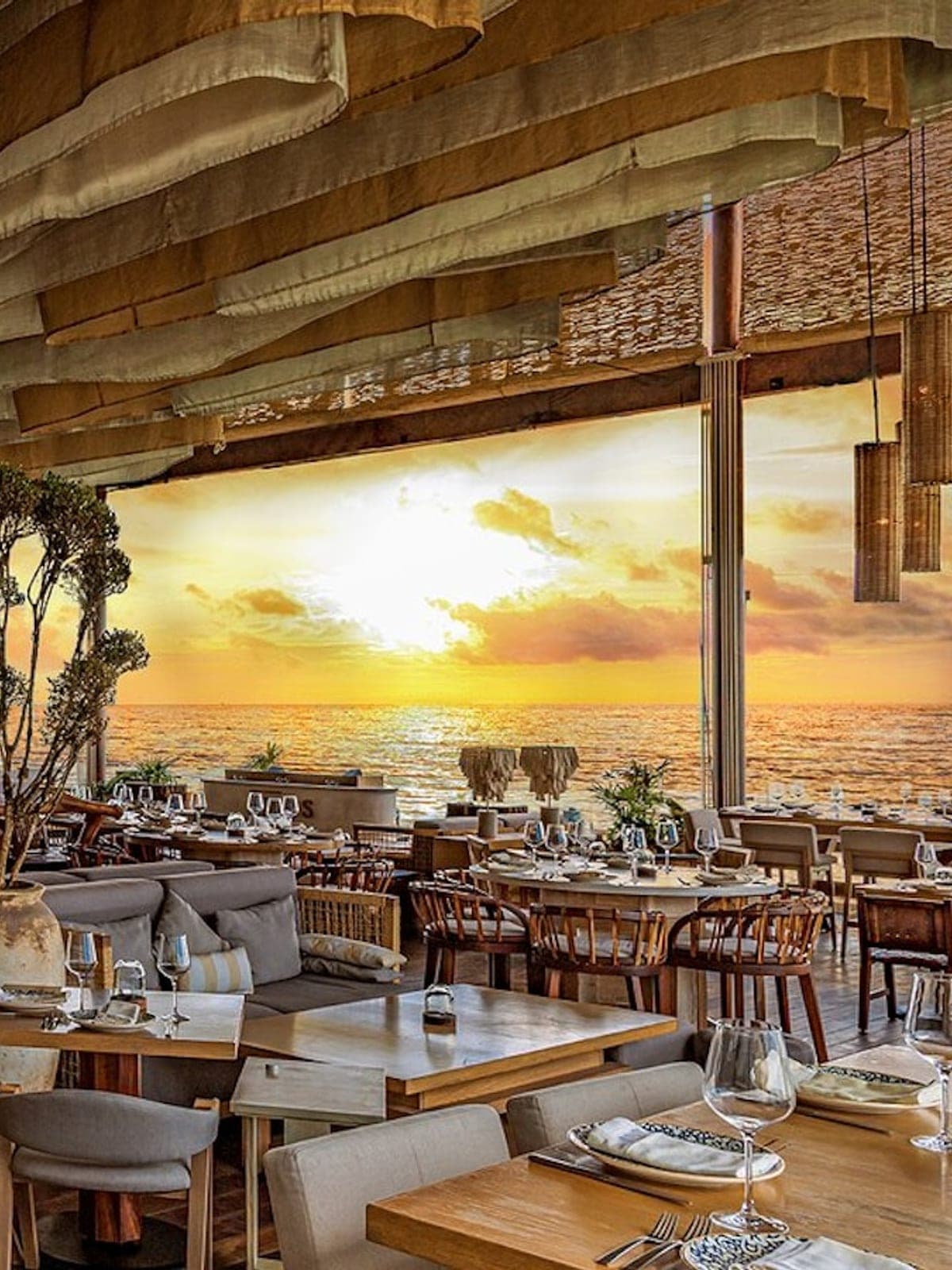 Ilios, cocina griega en Cancún: salón luminoso con mural de atardecer y columnas de madera.